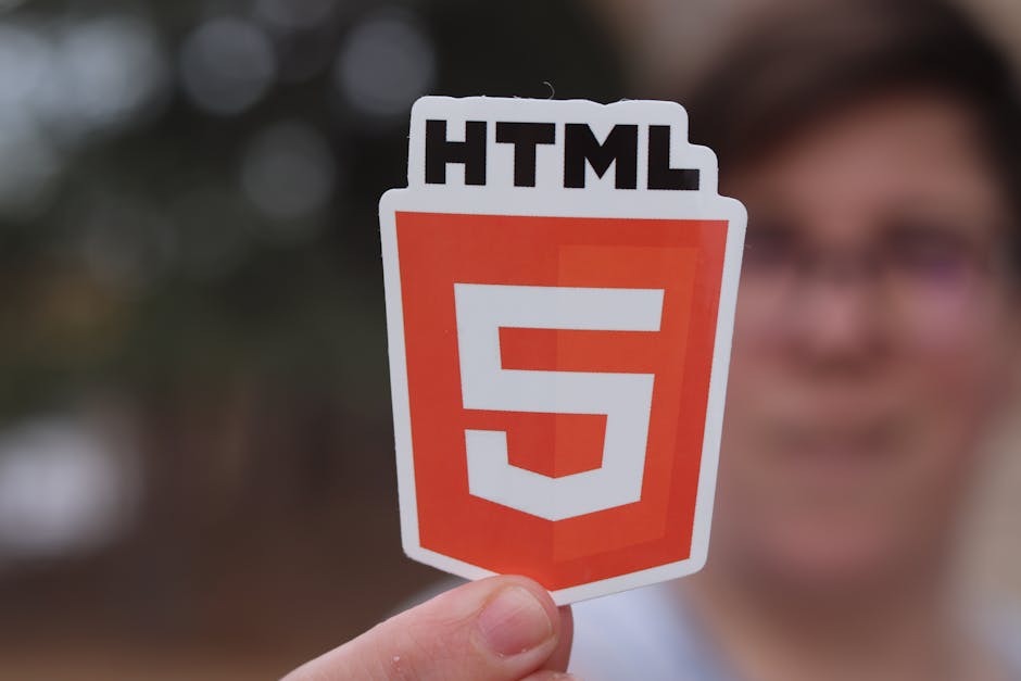 A person holding a sticker with the HTML5 logo, featuring a bold red shield shape with a large white number 5 and the word 'HTML' at the top. The individual is outdoors with a blurred background of tr